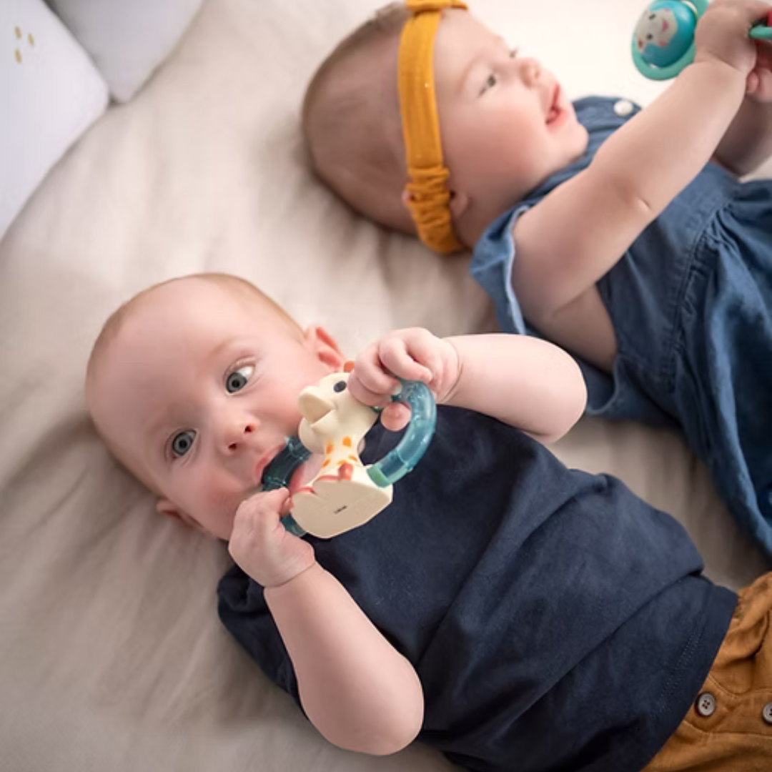 Two babies playing with Sophie La Girafe teething toys on a soft surface