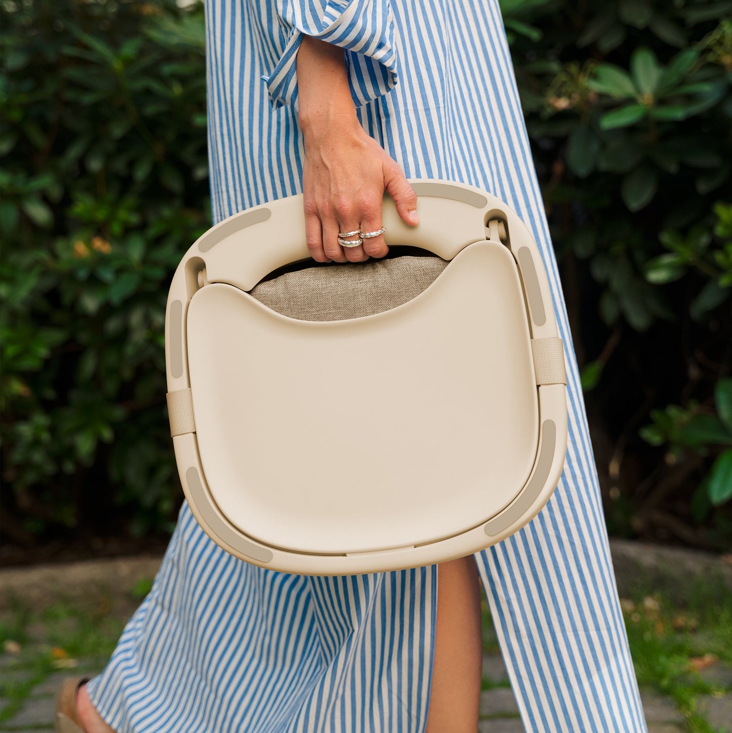 Person holding a beige portable highchair with a blurred green outdoor background