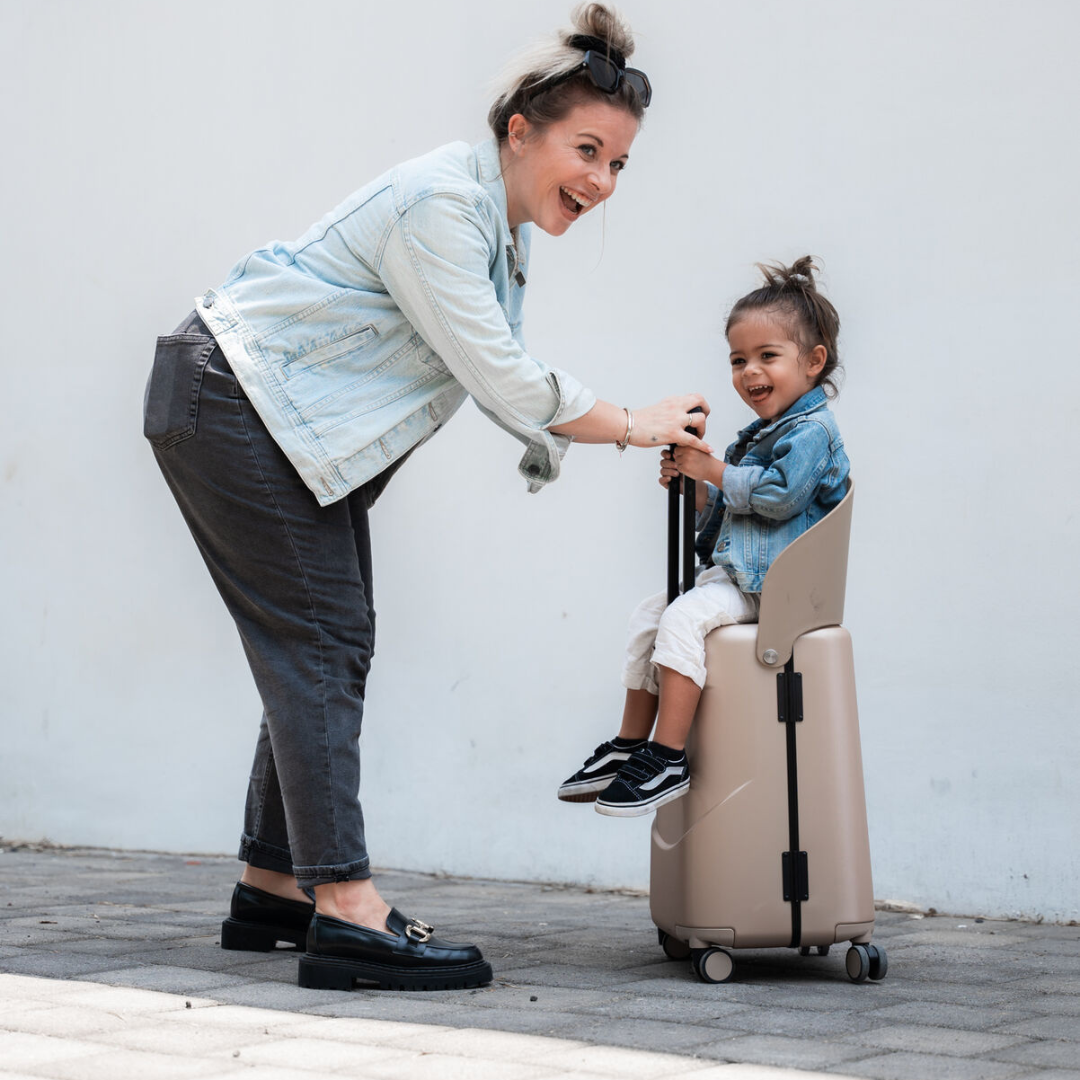 Woman and child playing with a beige suitcase against a white wall. Child shown sitting on suitcase seat attached. 