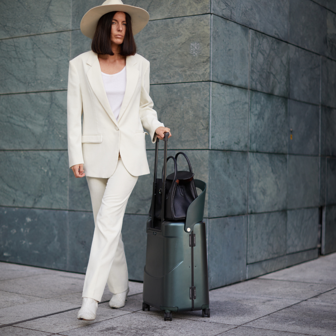 Woman in a white suit with a hat holding a green suitcase against a gray wall.