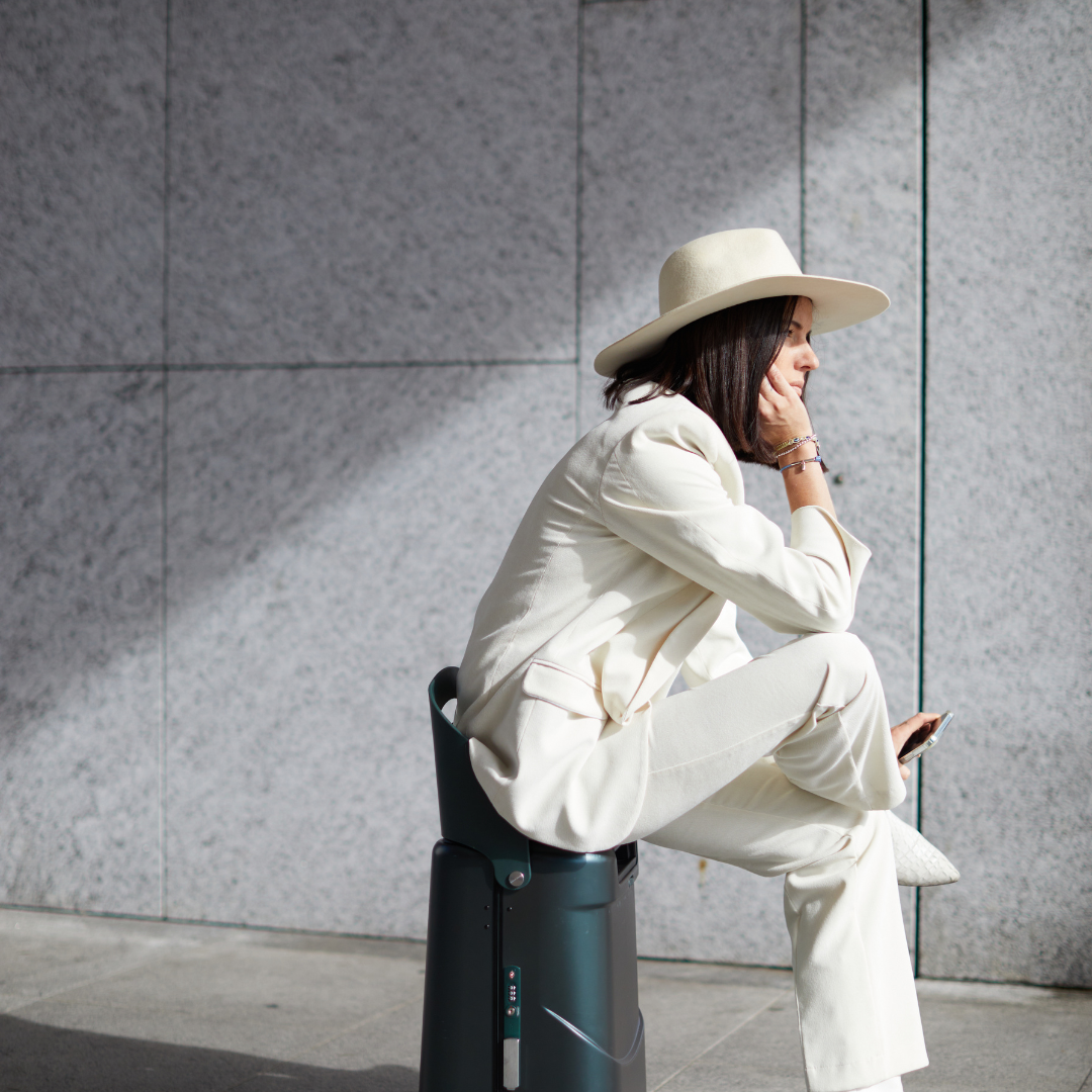 Person sitting on a green carry on suitcase against a grey wall wearing a white suit and beige hat.