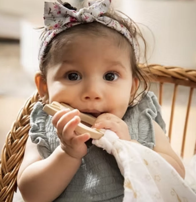 Baby with a wooden teething toy wearing a floral headband indoors.