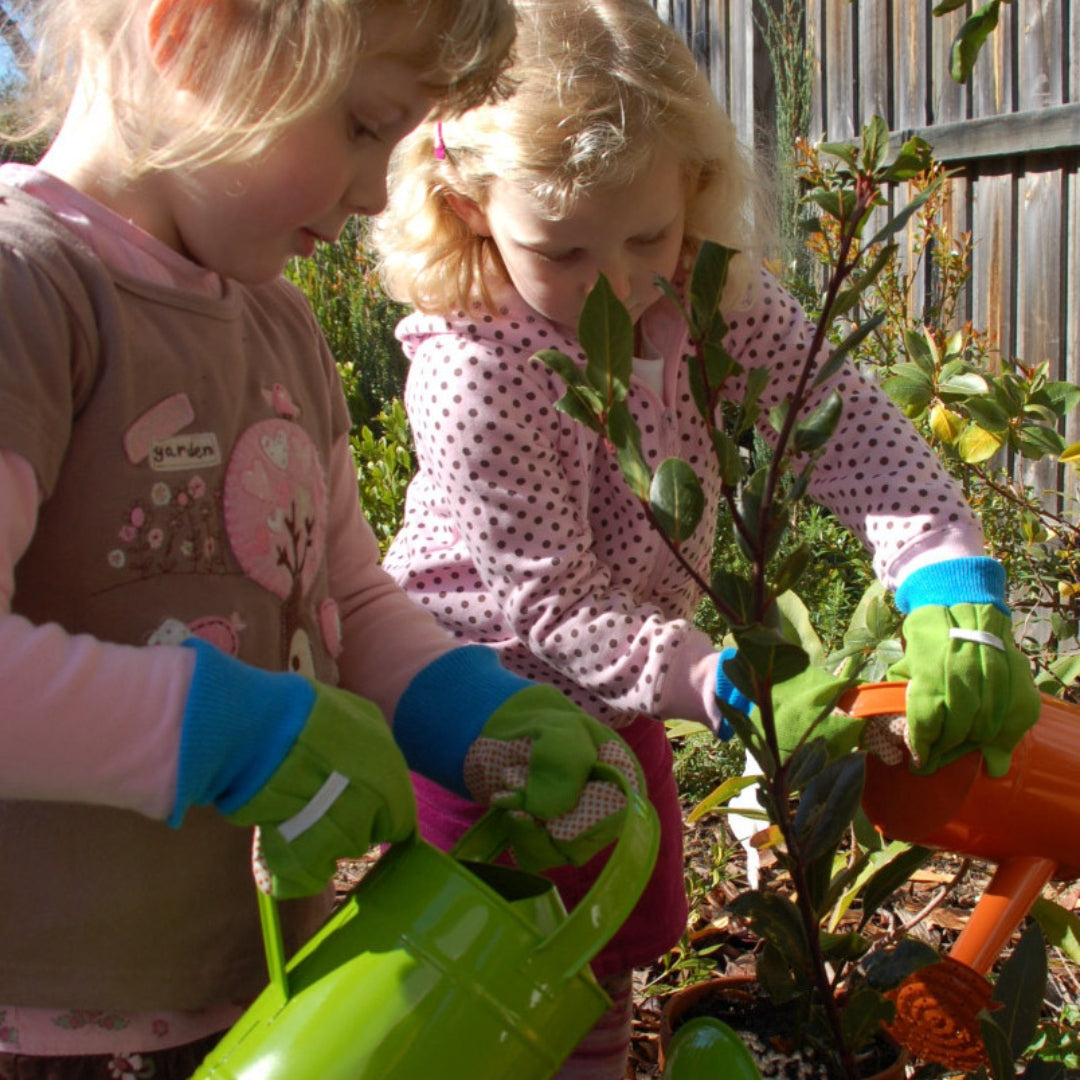 Two young girls in a garden, one watering a plant with a green watering can and the other holding an orange one with green gardening gloves. 